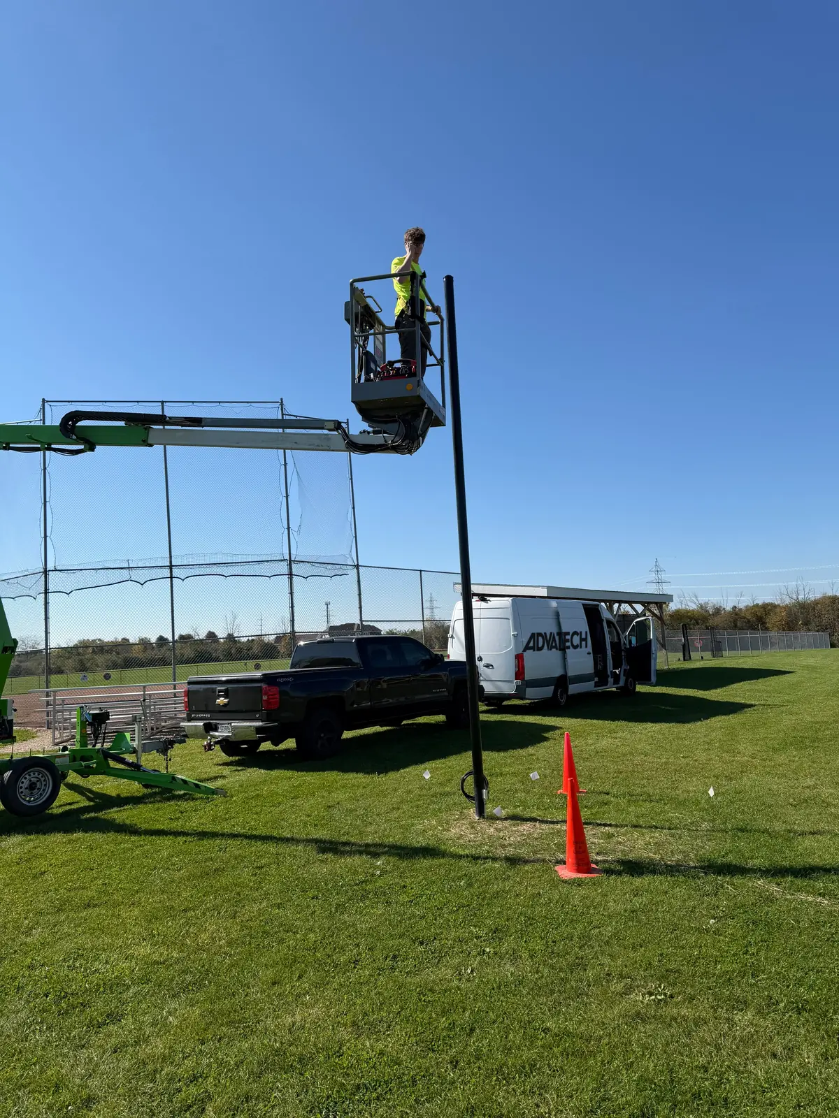 AdvaTech technician in a boom lift basket at the top of a camera pole beside a fenced ball field at a Mequon park