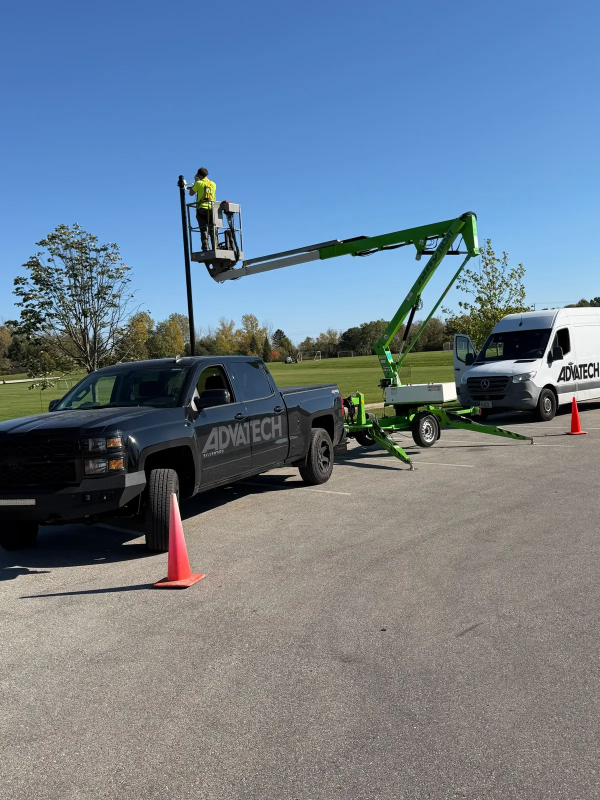 Towable boom lift raised at a camera pole in a Mequon park, with the AdvaTech Security van and pickup truck parked nearby and safety cones out